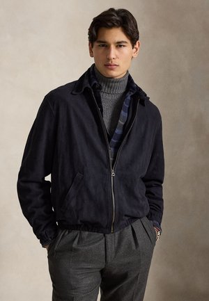 Jeune homme aux cheveux foncés portant une veste noire zippée par-dessus un col roulé gris et un pantalon gris, debout devant un fond neutre.