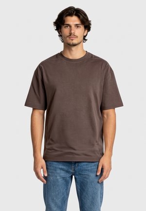 Młody mężczyzna o średniej długości ciemnych włosach nosi gładką brązową koszulkę z krótkim rękawem i niebieskie dżinsy, stojąc na jasnym tle.