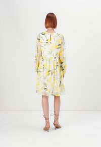 Blomstrete kjole med hvit bakgrunn, gule sitroner og grønne bladmønstre, lange ermer og samlet midje. Detalj med nøkkelhull bak.