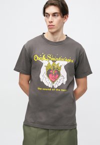Camiseta de algodón gris con un diseño gráfico de manos sosteniendo un corazón con llamas, acentuado con texto amarillo y rosa arriba y abajo.