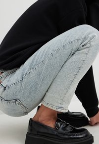 Jeans en denim bleu clair légèrement usés avec une coupe fuselée, présentant des décolorations subtiles et des coutures contrastées, assortis à des mocassins en cuir noir.