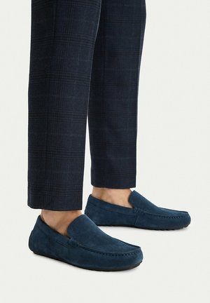 Granatowe zamszowe mokasyny noszone z ciemnymi, kratkowymi spodniami o kroju 7/8, odsłaniającymi gołe kostki na jednolitym jasnym tle.