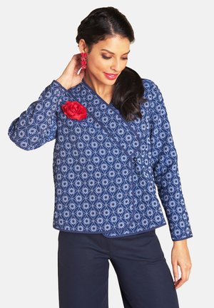 Frau trägt eine blaue Jacke mit Muster, einer roten Blütenbrosche und passenden roten Ohrringen, posiert mit einer Hand nahe dem Ohr.