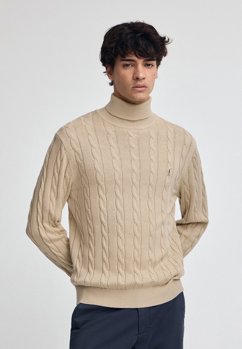 Beige stickad polotröja med ett kabelstickat mönster, ribbade muddar och nederkant, samt en liten logotyp på vänster bröst.