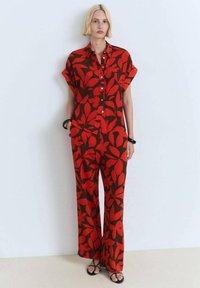 Red floral patterned shirt and wide-leg pants in a brown background; short sleeves, button-down front, and relaxed fit. Black sandals.