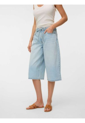 Žena nosí svetlomodré široké denim culottes so zaväzovaním v páse, biely tielkový top a hnedé ploché sandále.