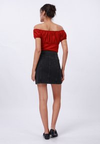 Femme se tenant de dos, portant un haut rouge à épaules dénudées, une jupe en denim noire, des boucles d'oreilles créoles et des chaussures plates noires sur un fond blanc.