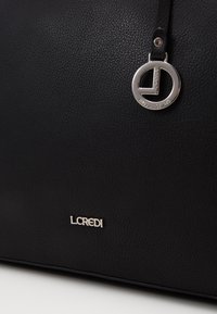 Détail d'un sac en cuir noir texturé avec un charm rond en métal avec le logo et le nom de marque L.CREDI en argent sur le devant.