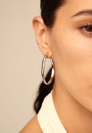 Primo piano dell'orecchio di una donna che indossa un orecchino a cerchio d'argento lucido e irregolare, su uno sfondo beige neutro.