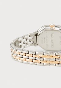 Montre en acier inoxydable argent et or avec un cadran rectangulaire. Elle présente une couronne en cristal et un bracelet à maillons avec des accents polis.