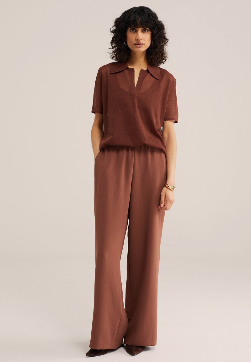 Femme aux cheveux bouclés foncés portant un haut marron à manches courtes avec col et un pantalon taille haute à jambes larges, debout, mains dans les poches.