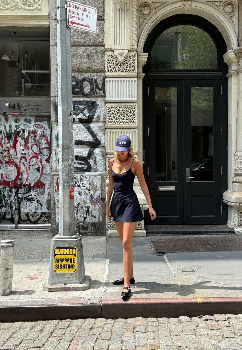 Robe noire sans manches avec jupe évasée, portée sur des jambes nues. Casquette noire avec le texte "NYC". Pavé en pierres et bâtiment couvert de graffitis en arrière-plan.