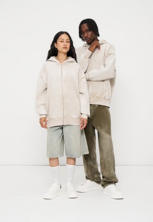 Zwei junge Erwachsene, die beige Kapuzen-Sweatshirts mit Reißverschluss tragen, stehen nebeneinander vor einem schlichten weißen Hintergrund.