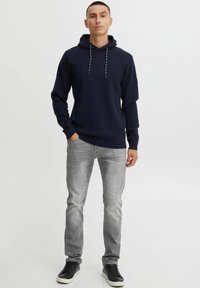 Marineblauwe hoodie met trekkoorden, ribbels aan de mouwen en onderkant, in combinatie met lichtgrijze jeans en zwarte sneakers, staand voor een witte achtergrond.