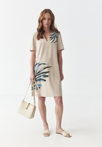 Abito beige con maniche corte e scollo a V, caratterizzato da una stampa di foglie blu e verdi. Il modello tiene una borsa beige chiaro e indossa sandali piatti.