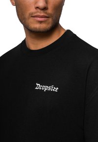 Sort sweatshirt med rund halsudskæring, prydet med hvid broderet tekst "Dropsize" på venstre brystområde. Materialet er en bomuldsblanding med en glat tekstur.