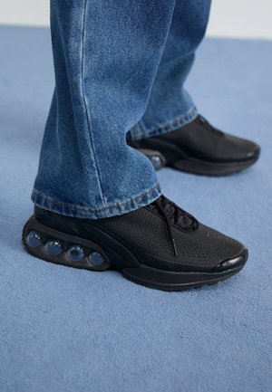 Baskets de sport noires avec tige en mesh, accents texturés et poches de coussin d'air visibles dans la semelle. Portées avec un jean en denim bleu.