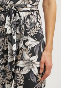 Tissu noir à motif floral avec des fleurs beiges et blanches noué à la taille, avec la main droite d'une personne reposant à côté.