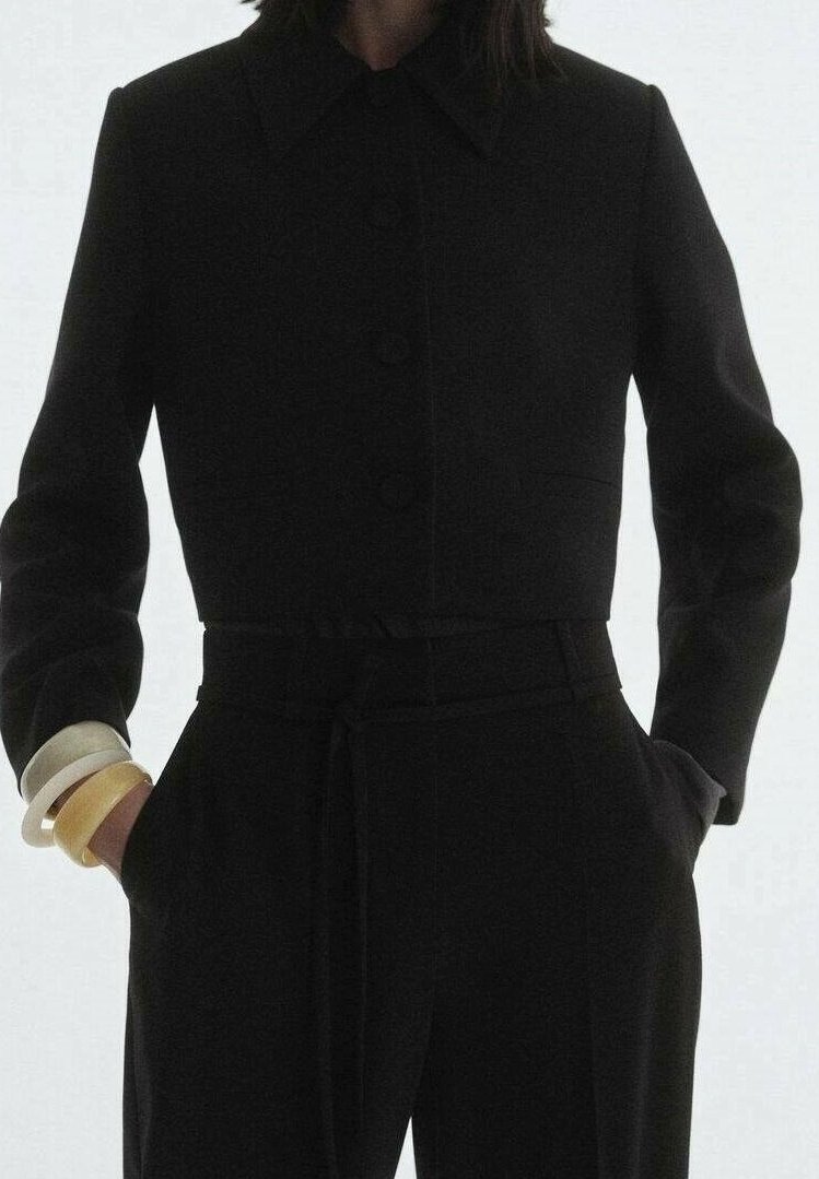 Personne portant un blazer noir cintré et un pantalon noir taille haute, les mains dans les poches, accessoirisée avec des bracelets beiges et blancs.