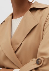 Beige trenssitakki, jossa on lovikaulus, suuret napit ja sileä pinta, yhdistetty vaaleaan, resorilliseen neulepaitaan alla.