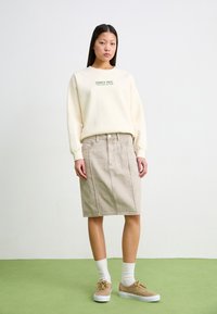 Cream sweatshirt with green printed text, paired with a beige denim skirt. Beige sneakers and white socks complete the outfit. Green background.