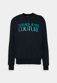 Svart sweatshirt med ribbad rundad halsringning och långa ärmar. Framsidan visar "VERSACE JEANS COUTURE" i iriserande bokstäver.