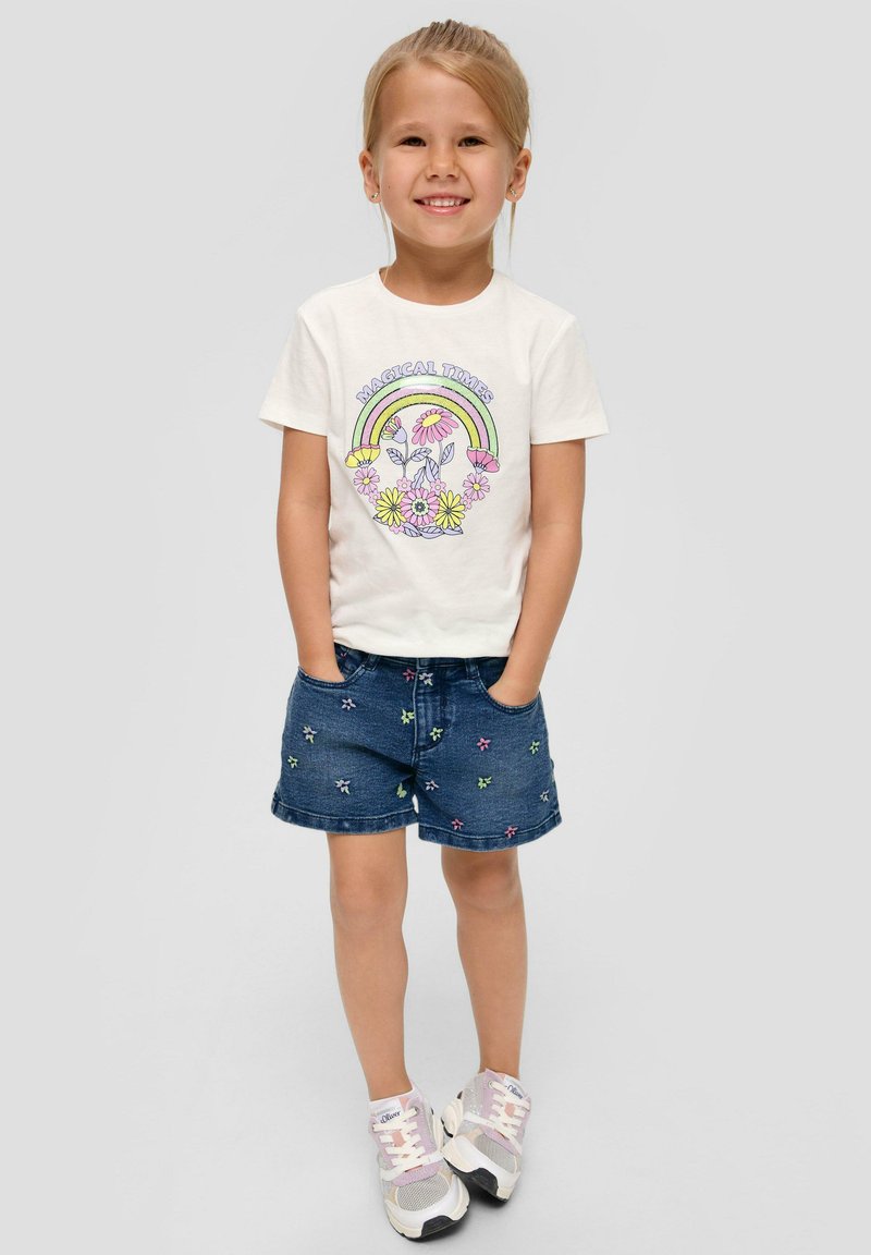 Weißes Baumwoll-T-Shirt mit "MAGICAL TIMES" Grafik und Regenbogen-Design; blaue Denim-Shorts mit mehrfarbiger Blumenstickerei; graue Sneakers.