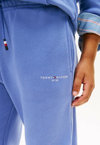 Lyseblå sweatshirt-stof med en glat tekstur, der har et hvidt "TOMMY HILFIGER" logo og en snoretræk med tricolor akcent.