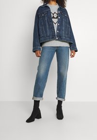 Veste en denim foncé avec un design court, pull gris en dessous, jean bleu clair retroussé, et bottines noires à talons carrés.