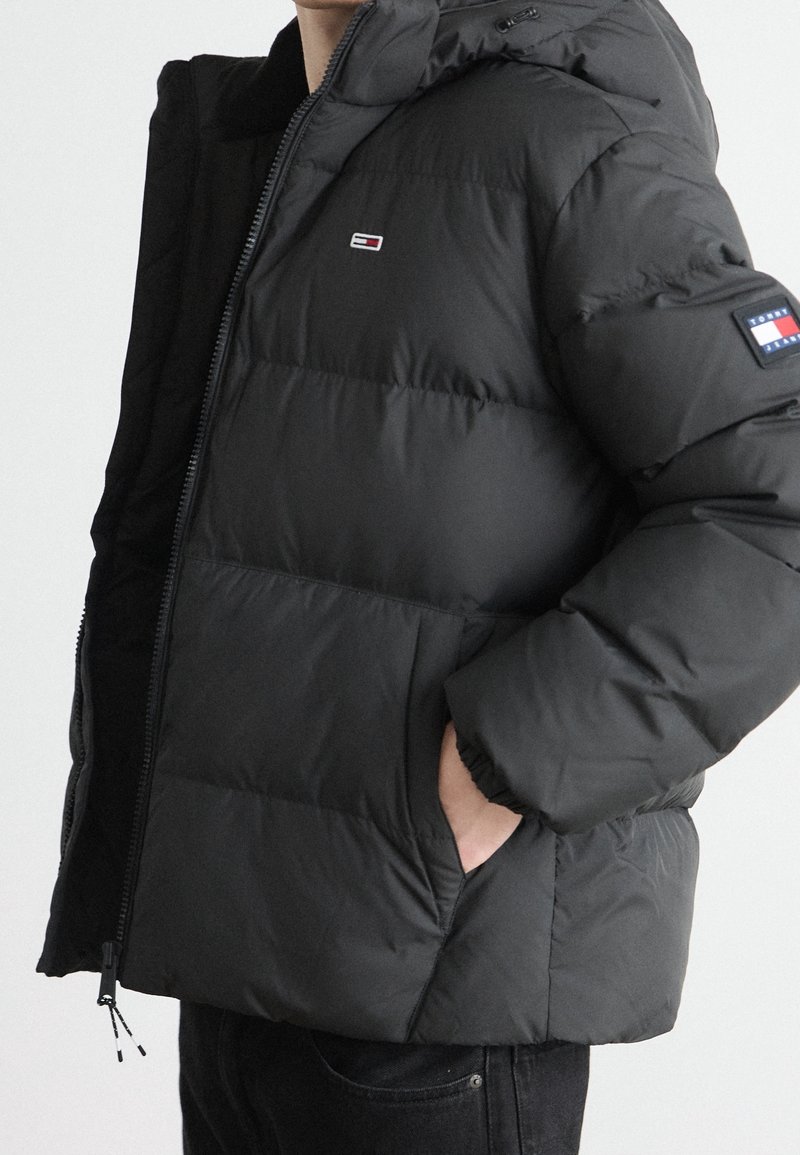 Personne portant une veste matelassée noire avec une capuche, main dans la poche, avec un petit logo rectangulaire sur la poitrine et un patch sur la manche gauche.