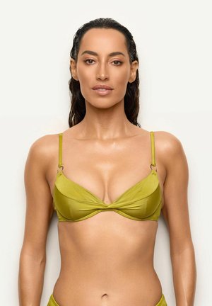 Mujer con el pelo mojado que lleva un top de bikini verde oliva, mirando hacia delante sobre un fondo blanco liso.