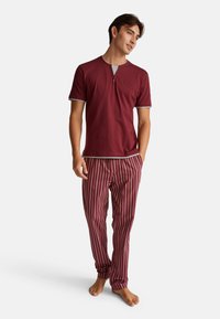 Man in een bordeauxrode short-sleeve henley shirt en bijpassende gestreepte bordeauxrode pyjamabroek, staand op blote voeten tegen een witte achtergrond.