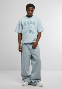 Tricou oversized de culoare albastru deschis cu imprimeu "KARL KANI NYC USA" în albastru închis, asortat cu blugi lejeri de culoare albastru deschis și adidași albi.
