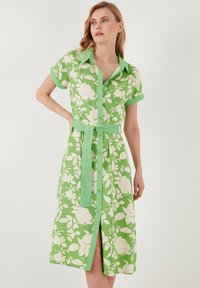 Grünes Blumenkleid mit weißen botanischen Mustern, kurzen umgeschlagenen Ärmeln, Knopfleiste und einem passenden Gürtel in der Taille. Glatter Stoff.
