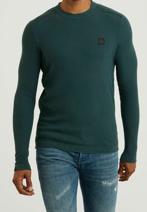Homme portant une chemise à manches longues texturée vert foncé avec un petit logo sur la poitrine et un jean bleu délavé.