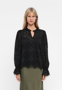 Femme portant un haut en dentelle noire à manches longues avec des motifs floraux et des poignets volants, associé à une jupe vert olive sur fond blanc.