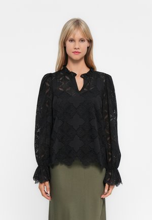 Donna che indossa una blusa nera in pizzo con maniche lunghe e motivi floreali, con polsini a ruffles, abbinata a una gonna verde oliva su uno sfondo bianco.