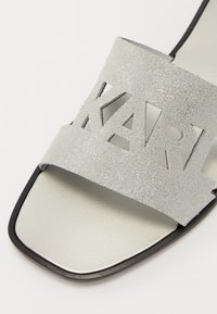 Sandalias de purpurina plateada con una correa ancha que presenta letras recortadas que deletrean "KARL." La suela es de color gris claro con un brillo sutil. Diseño de punta cuadrada.