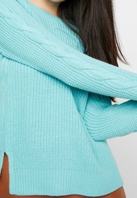 Pull bleu clair en maille avec un motif torsadé sur les manches, doté d'un ourlet côtelé et de fentes latérales pour une coupe décontractée.
