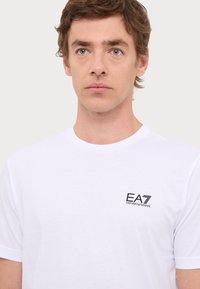 Fehér pamut póló, kerek nyakkivágással. A bal mellen fekete EA7 Emporio Armani logó látható. Egyszerű, rövid ujjú dizájn.