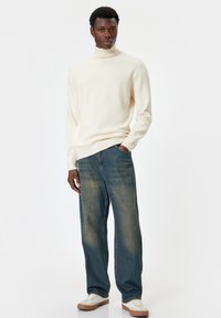 Kremowy sweter z wysokim kołnierzem, szerokie, wyblakłe niebieskie dżinsy i białe sneakersy z brązowymi podeszwami. Gładka faktura, luźny krój i swobodny styl.