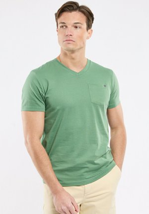 Homme portant un T-shirt vert à encolure en V avec une poche poitrine et un pantalon beige, debout avec une main dans la poche, regardant sur le côté.