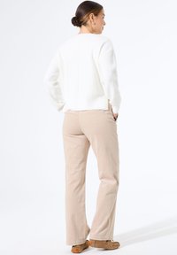 Vrouw met bruin haar in een knot, gekleed in een witte trui, beige pantalons en bruine moccasin schoenen, staand met handen in de zakken en van zich af gekeerd.