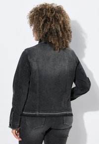 Mustapekka denim-takki, jossa on tiukka muotoilu, näkyvät ompeleet ja hienovarainen haalistus. Varusteena korkea kaulus ja nappi- sekä hihansuut.