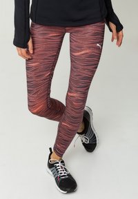Osoba ubrana w legginsy Puma w czarno-różowy wzór, czarne i szare buty sportowe oraz czarną bluzę z długim rękawem, stojąca na jasnym tle.