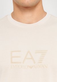 Šviesiai kreminė marškinėlių suapvalinta apykaklė pagaminta iš minkštos medžiagos, centre iškeltas logotipas "EA7 EMPORIO ARMANI", su tekstūruotais raštais.