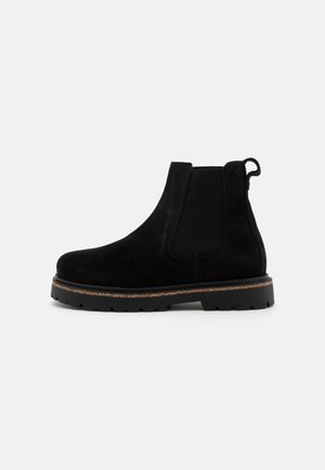 Zwarte suède Chelsea-boots met elastische zijpanelen, bevestigingslus en textuur rubberen zool. Heeft een laag profiel en een contrasterende bruine middenzool.