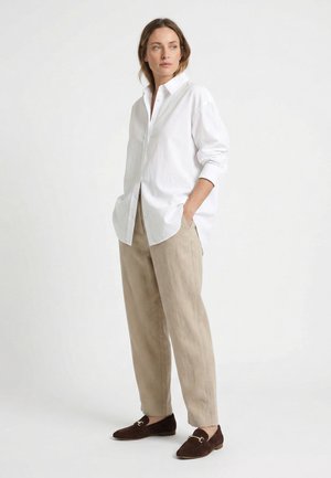 Frau steht mit Händen in den Taschen, trägt ein lockeres weißes Hemd mit Knöpfen, beige weite Hosen und dunkelbraune Loafers vor weißem Hintergrund.