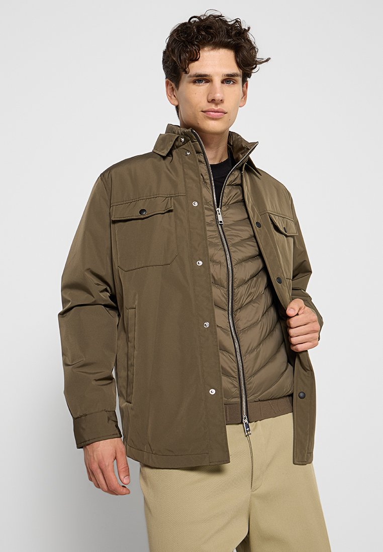 Veste vert olive avec deux poches poitrine, détails de boutons et une couche intérieure zippée. Tissu léger et coupe décontractée.