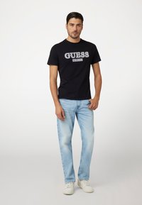 Juodas medvilnės marškinėliai su balta „GUESS EST.1981“ logo, derinami su šviesiai mėlynomis džinsinėmis kelnėmis. Modelis vilki baltus sportbačius.